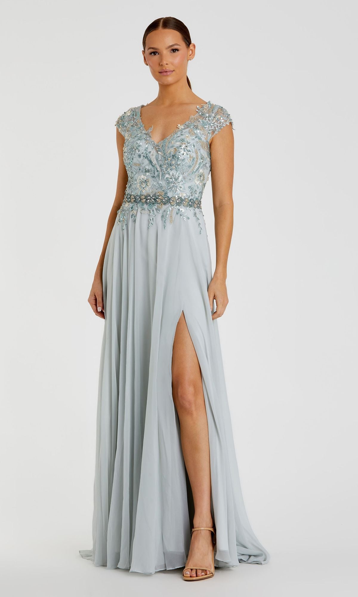 A woman stands against a plain background wearing the Mac Duggal 79365 cap-sleeve long formal dress in light blue, featuring a flowy skirt, embellished bodice with floral embroidery, cap sleeves, and a thigh-high slit.